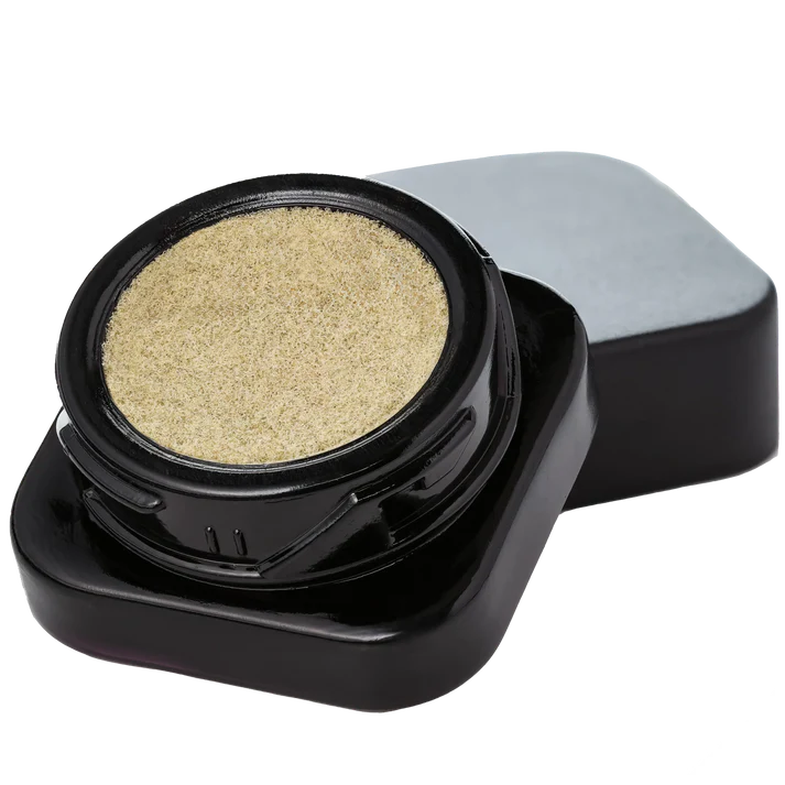 A small round cosmetic jar filled with a shimmering light gold cream or powder, shown open with the black lid resting behind it on a plain white background.
